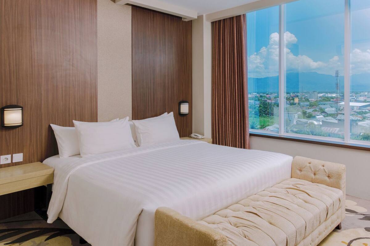Une chambre d’hôtel avec un grand lit, une table de chevet, une chaise et une vue sur le paysage urbain, visible à travers une grande fenêtre.