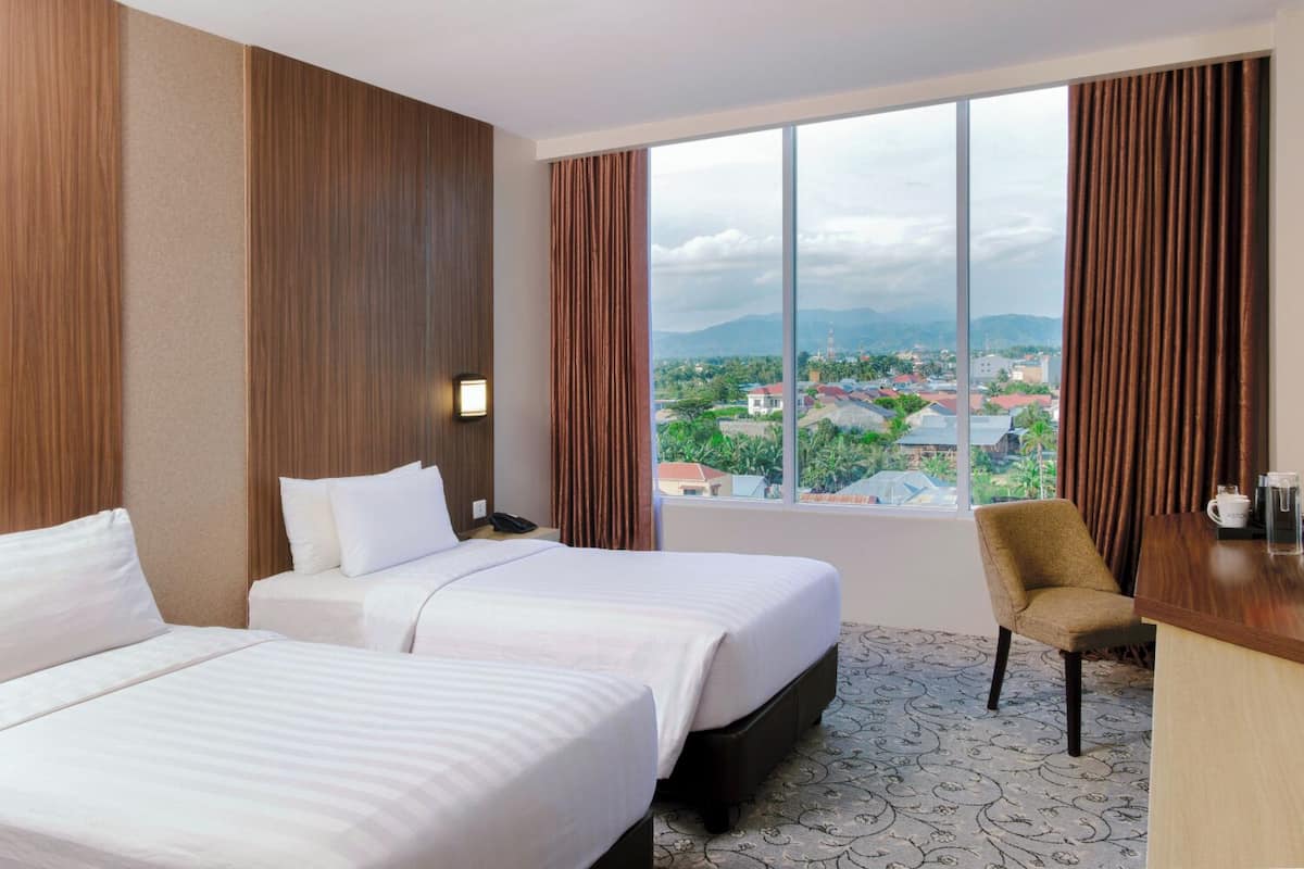 Une chambre d’hôtel avec deux lits, un bureau et une chaise. Une grande fenêtre offre une vue sur le paysage urbain et les montagnes.
