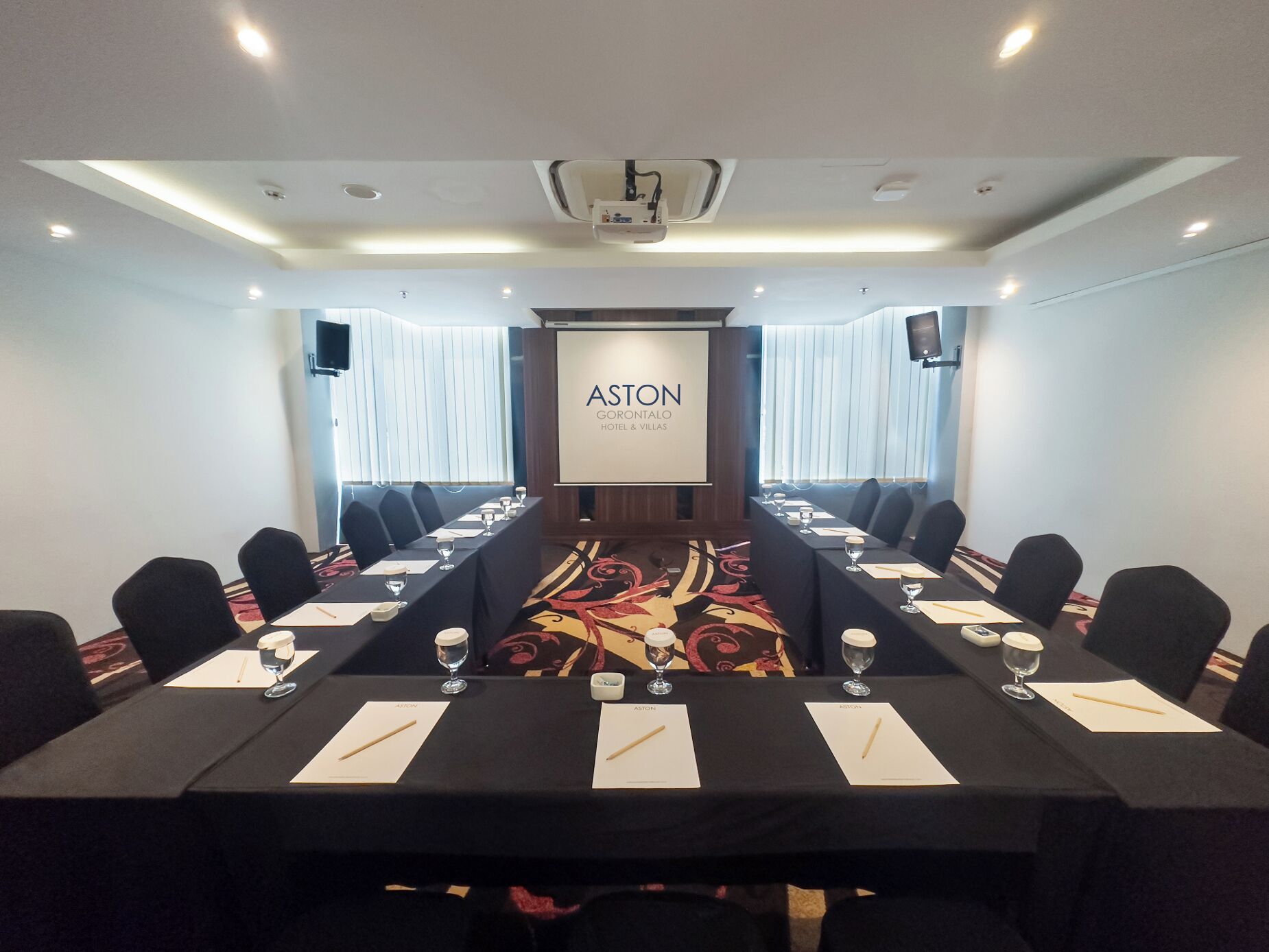 Ein Konferenzraum mit einem langen Tisch, Stühlen und einem Bildschirm, der das Logo "ASTON GORONTALO HOTEL & VILLAS" zeigt.