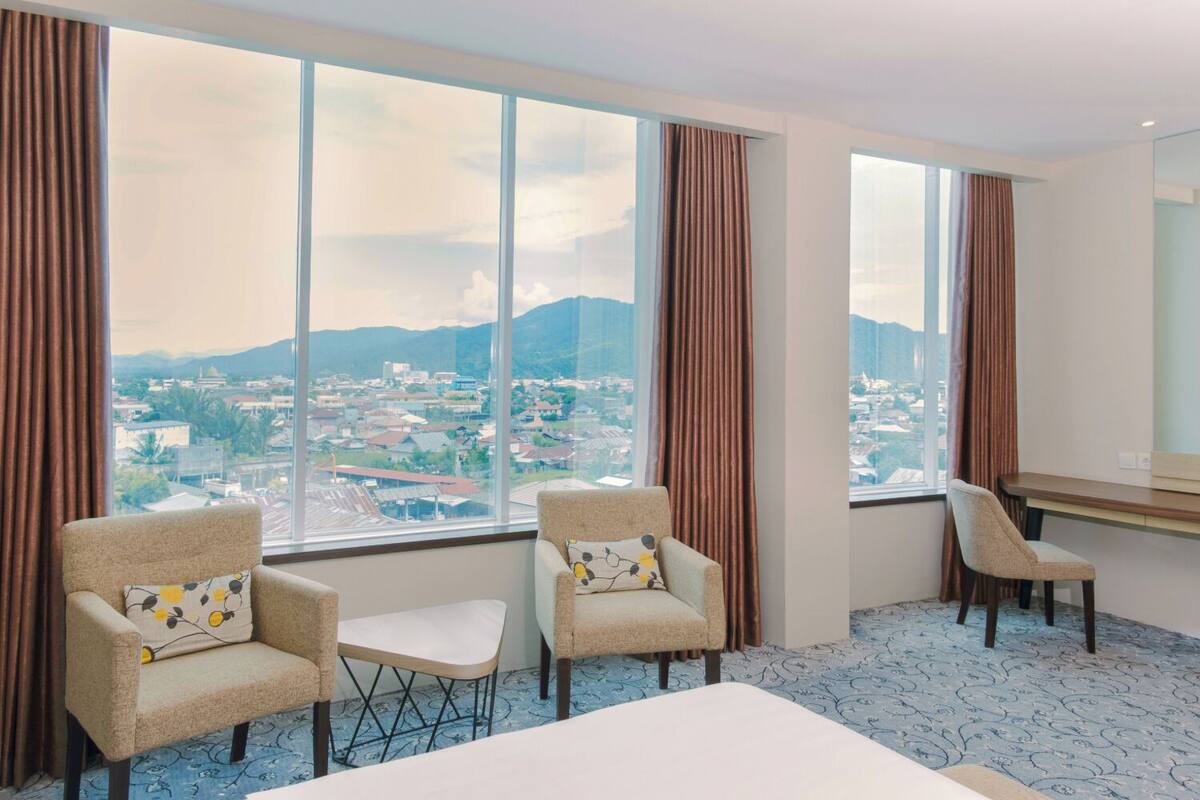 Une chambre d’hôtel avec un grand lit, deux fauteuils, une petite table et un bureau. La chambre dispose de grandes fenêtres offrant une vue sur le paysage urbain et les montagnes.
