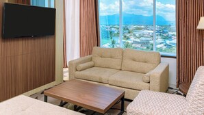 A hotel room with a large window offering a city view, a flat-screen TV mounted on the wall, a beige sofa, a wooden coffee table, and a patterned ottoman. - Aston Gorontalo Hotel & Convention Center (Gorontalo)