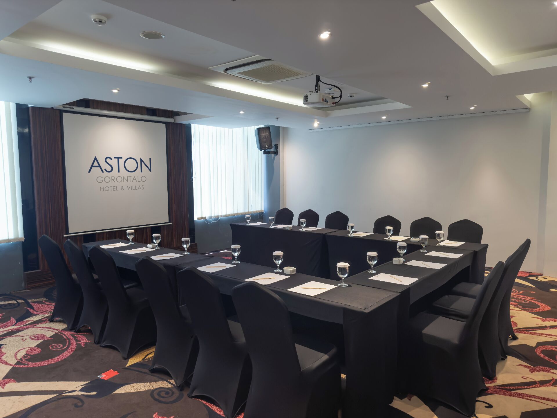 Une salle de conférence avec une longue table, des chaises noires et un écran affichant le texte « ASTON GORONTALO HOTEL & VILLAS ».