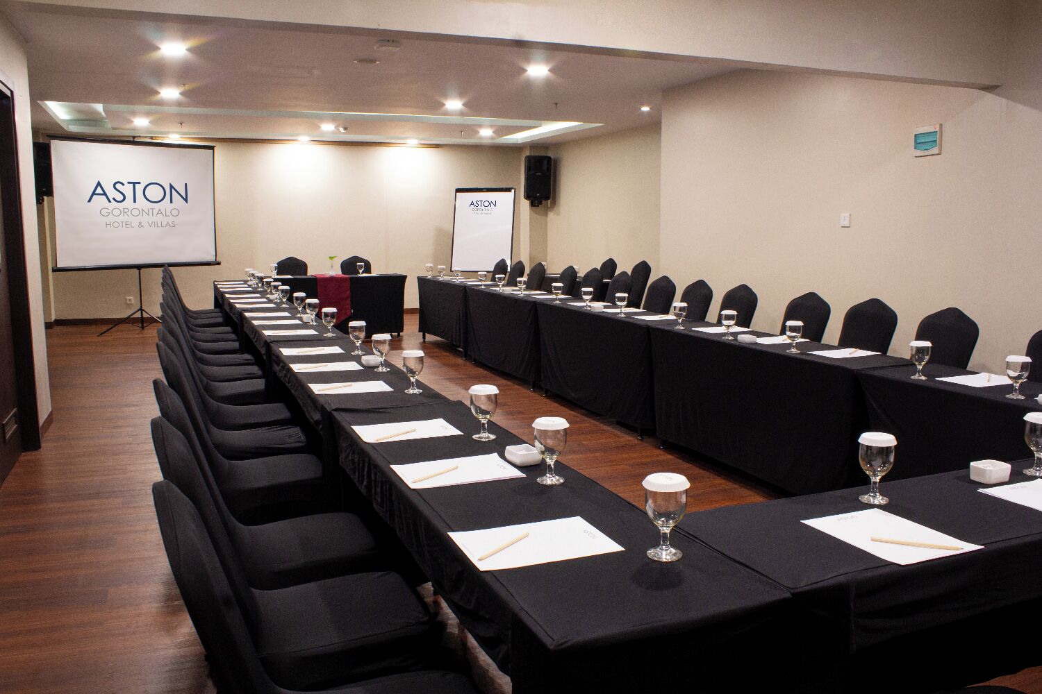 A conference room with long tables, black chairs, and a projector screen displaying the logo of Aston Hotel & Villas.
