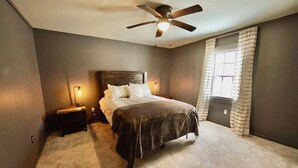 Un dormitorio con cama, mesita de noche, ventilador de techo y ventana con cortinas.
