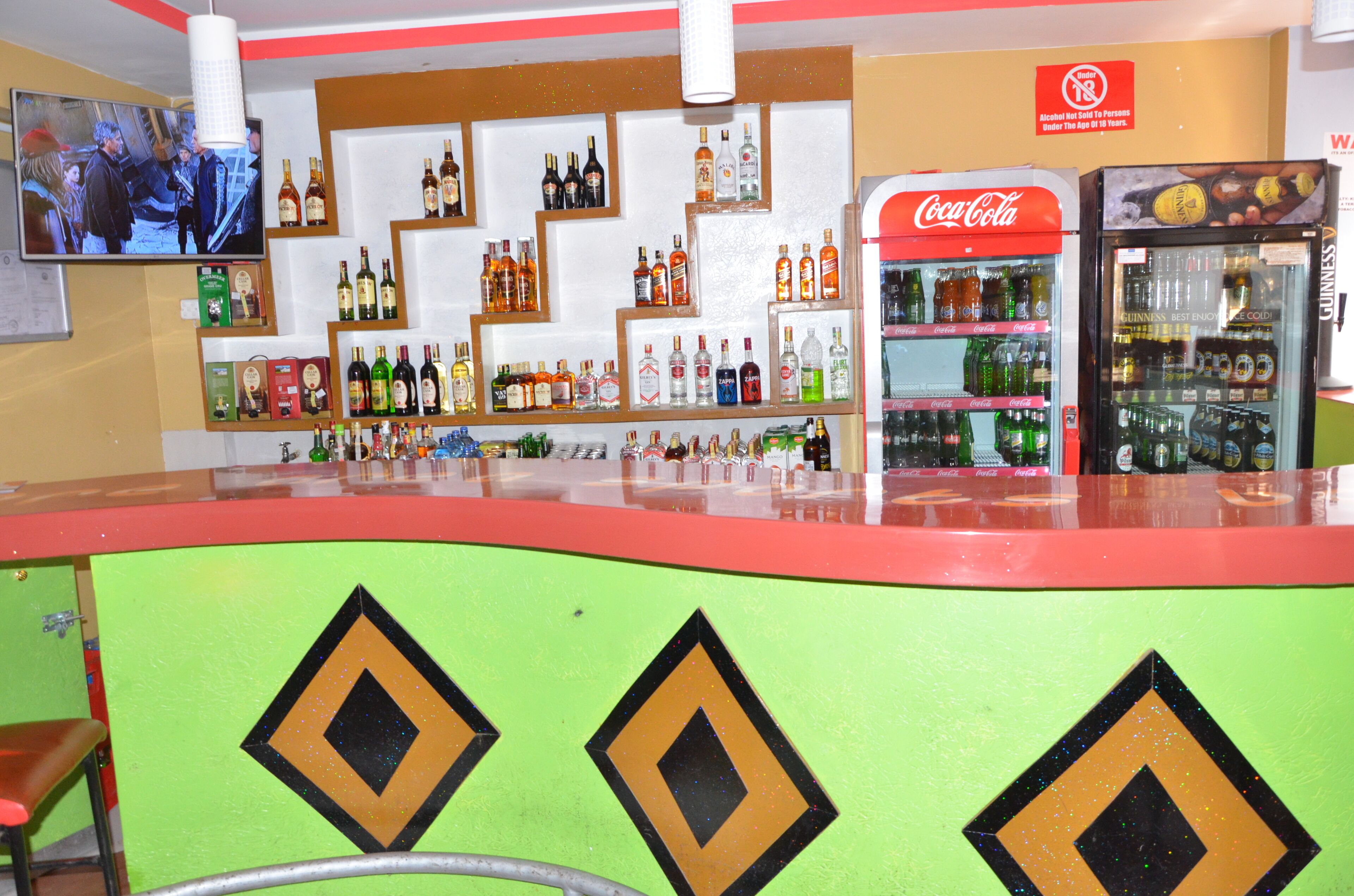 A bar area with shelves of liquor bottles, a Coca-Cola refrigerator, and a television screen.