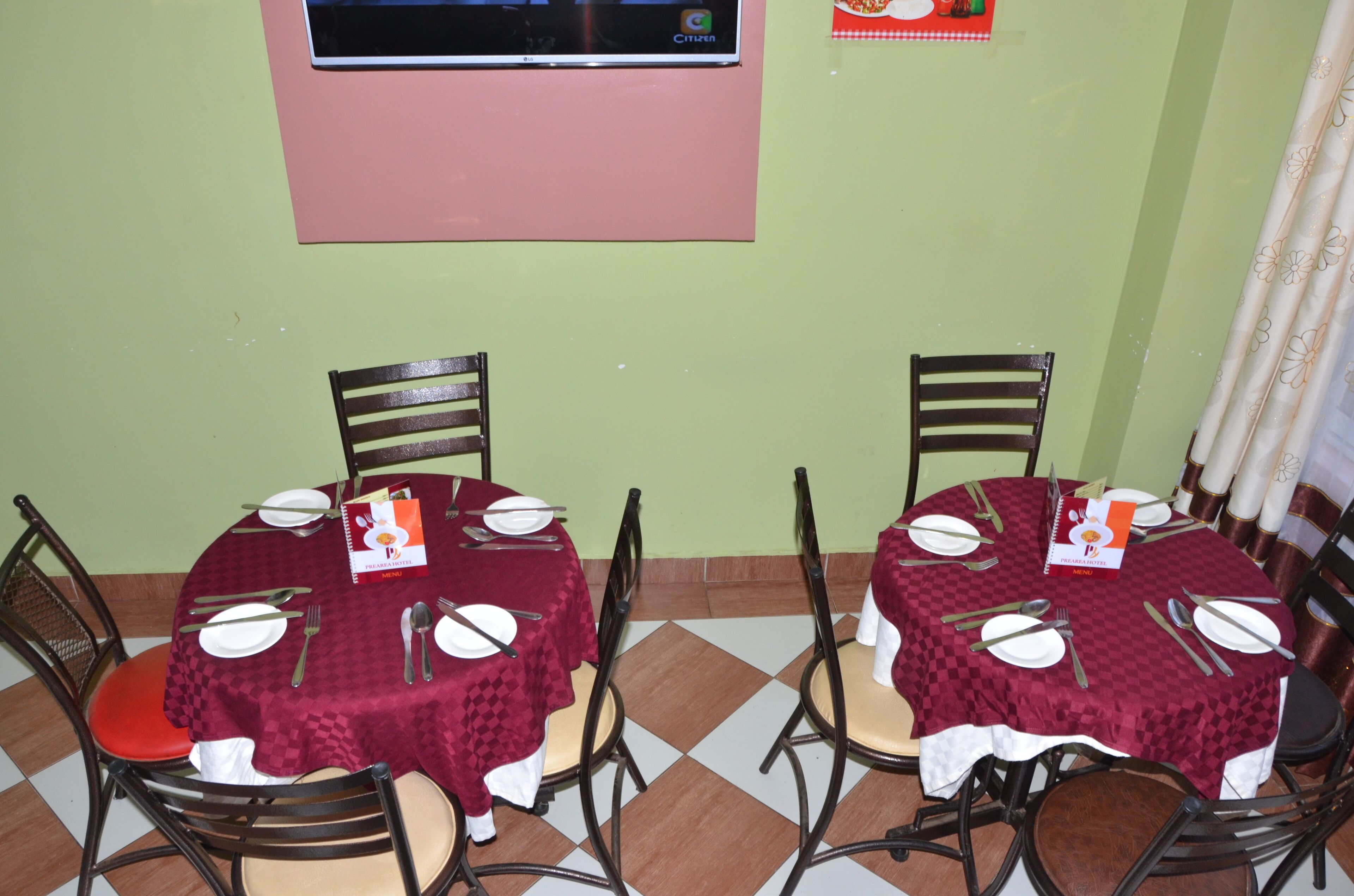 Un comedor con dos mesas redondas, cada una con platos blancos, cubiertos y servilletas. Las mesas están cubiertas con manteles color burdeos, y hay sillas alrededor. Un televisor está montado en la pared sobre las mesas.