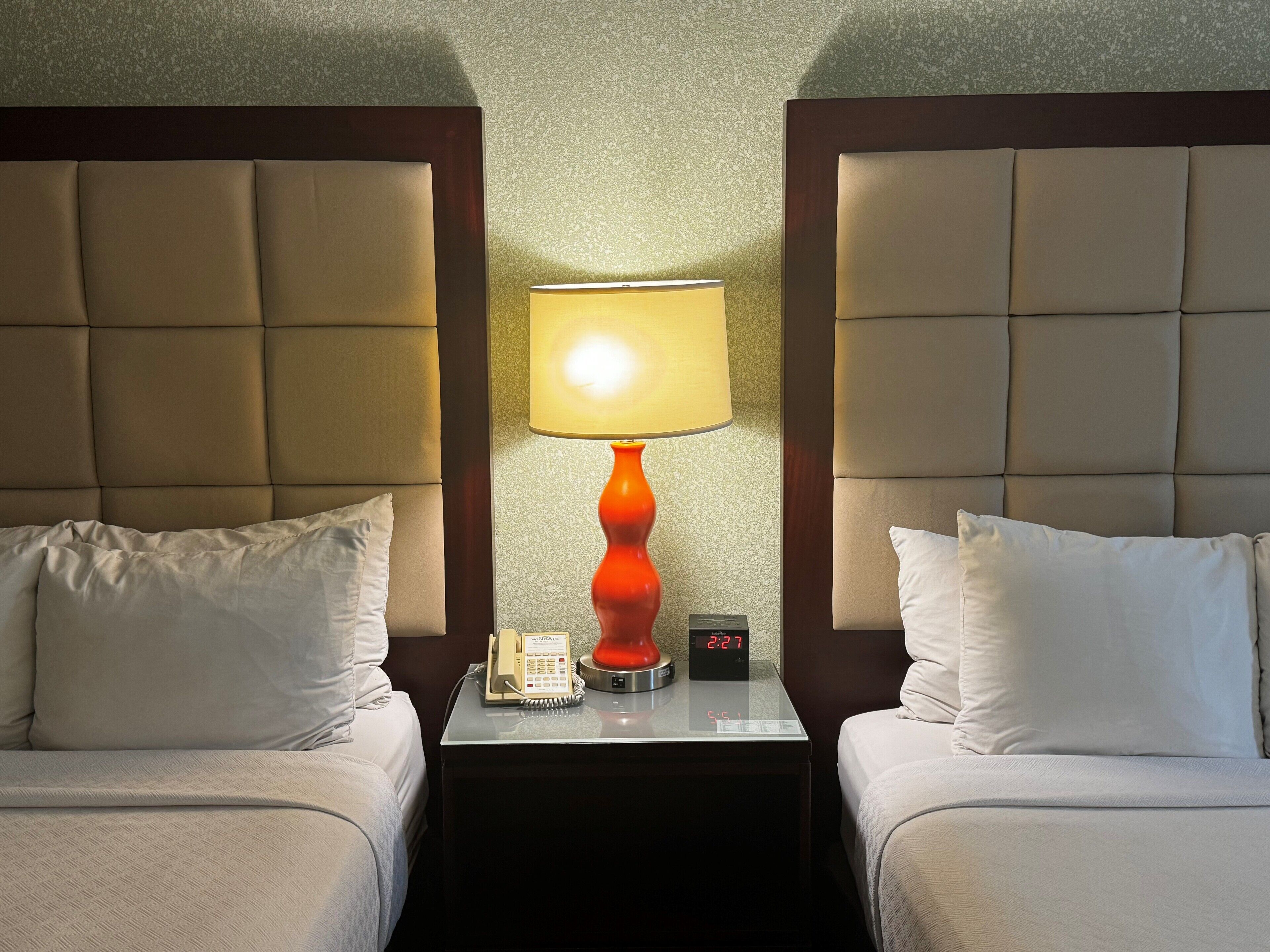A hotel room with two beds, a nightstand with a lamp, a phone, and a digital clock.