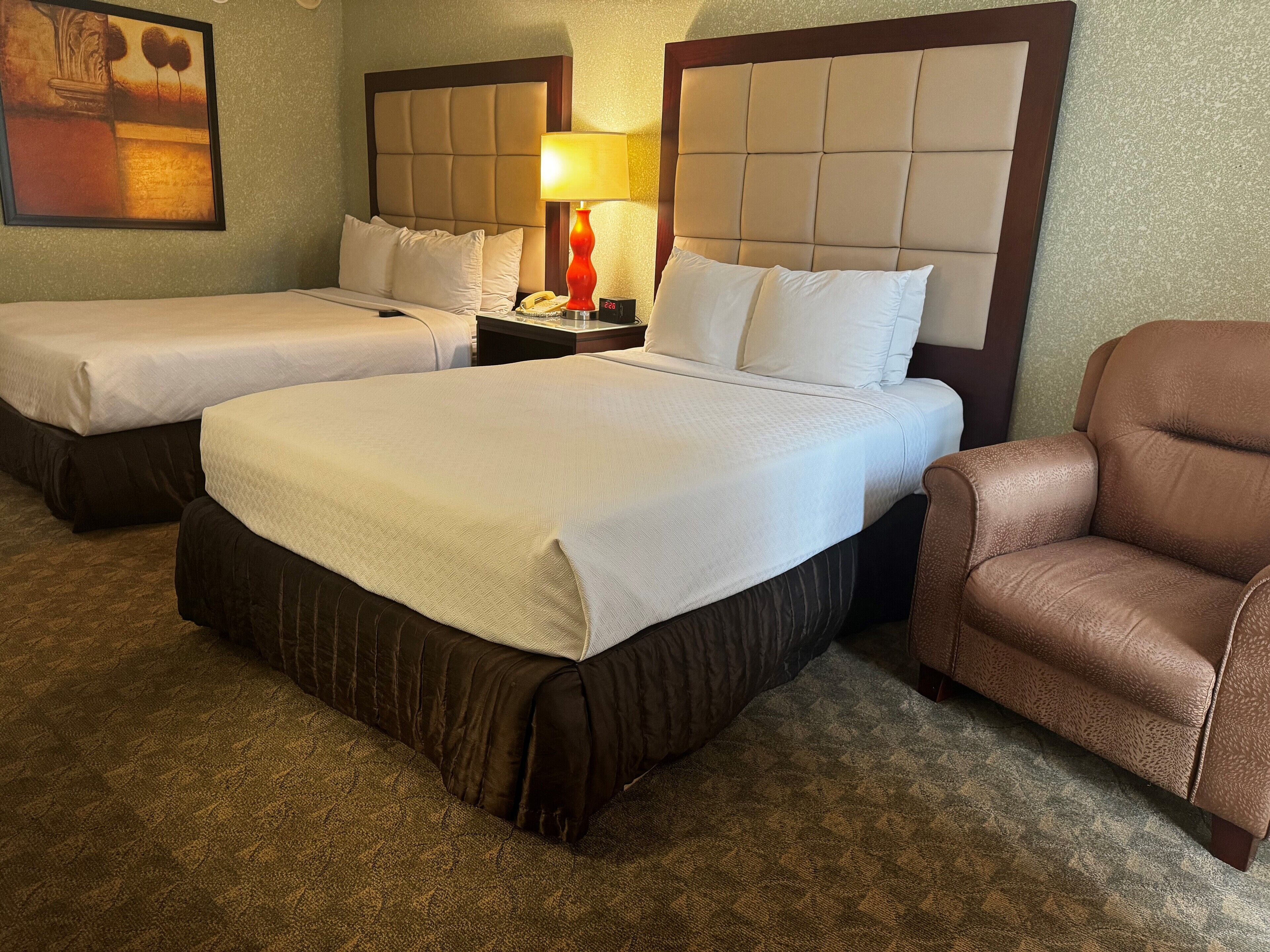 A hotel room with two beds, a nightstand with a lamp, a brown leather armchair, and a framed picture on the wall.