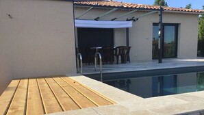 Een zwembad met een houten terras, stenen traptreden en een overdekt zitgedeelte met stoelen en een tafel.