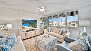 A living room with a large TV, a ceiling fan, and a view of the ocean through the windows. - Loggerhead Cay 274: Beautiful Condo on the Beach! (Sanibel)