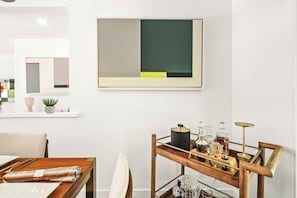 Een modern interieur met een minimalistisch design, met een houten eettafel, een metalen bijzettafeltje met glazen, en een groot abstract schilderij aan de muur.