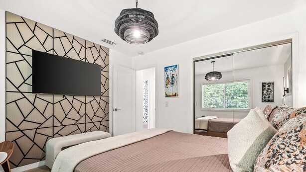 Ein modernes Schlafzimmer mit einem großen Bett, einem flachbildfernseher an einer Wand mit geometrischem Muster, einem großen Spiegel und einer Deckenleuchte.
