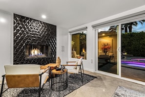 Een moderne woonkamer met een geometrisch wandontwerp, een open haard en een zithoek met uitzicht op een zwembad en buitenruimte.