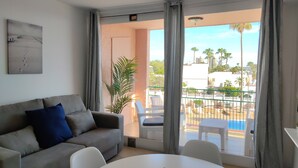 Ein Wohnzimmer mit grauem Sofa, einem kleinen Tisch und einem Balkon mit Blick auf Palmen und Gebäude.