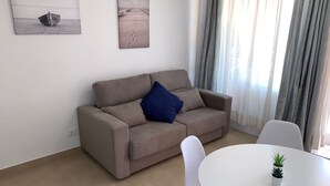 Ein modernes Wohnzimmer mit einem beigefarbenen Sofa, einem blauen Kissen, einem weißen runden Tisch mit zwei Blumentöpfen und Kunstwerken an den Wänden.