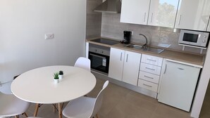 Eine kompakte Küche mit weißen Schränken, einem eingebauten Backofen, einer Mikrowelle und einer Kaffeemaschine. Ein runder Tisch mit Stühlen und ein kleiner Topfpflanze stehen dort.
