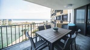 A balcony with a wooden table and chairs, overlooking a beach and a tennis court.