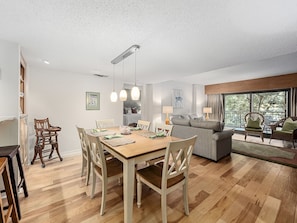 A dining area with a wooden table and chairs, a sofa, and a view of the outdoors through large windows.