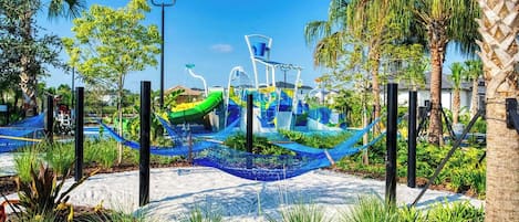 A playground with slides and climbing structures, surrounded by palm trees and a blue netted area.