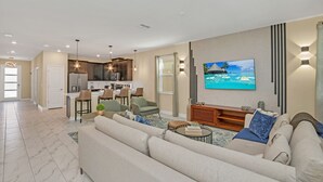Una sala moderna con un sofá grande, una mesa de centro y un televisor montado en la pared. Se ve un área de cocina con una mesa de comedor y sillas al fondo.