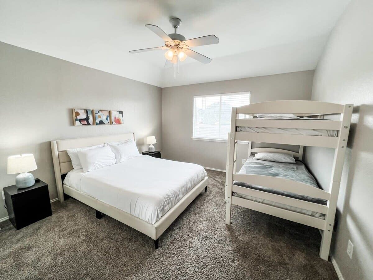 A bedroom with a bunk bed, a single bed, a ceiling fan, a nightstand with a lamp, and a window with blinds.