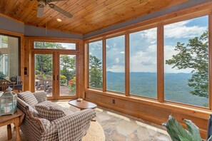 A cozy room with a checkered sofa, a stone floor, and large windows offering a view of a forest and mountains. - Spectacular Mountain retreat- First time this home offered as a rental (Highlands)
