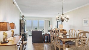 A spacious living area with a dining table, chairs, and a chandelier. There is a view of the ocean through large windows. - Shorewood 403- Oceanfront 4th Floor Condo (Hilton Head Island)