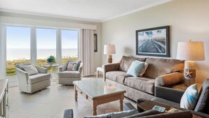 A spacious living room with a large window offering a view of the beach, a brown leather sofa, a glass coffee table, and two armchairs. - Shorewood 403- Oceanfront 4th Floor Condo (Hilton Head Island)