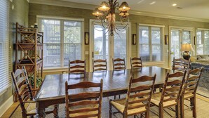 Uma sala de jantar com uma grande mesa de madeira rodeada por cadeiras, um candelabro e amplas janelas com persianas.
