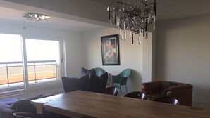 Un coin repas avec une table et des chaises en bois, un lustre moderne et une vue sur la plage grâce à de grandes fenêtres.