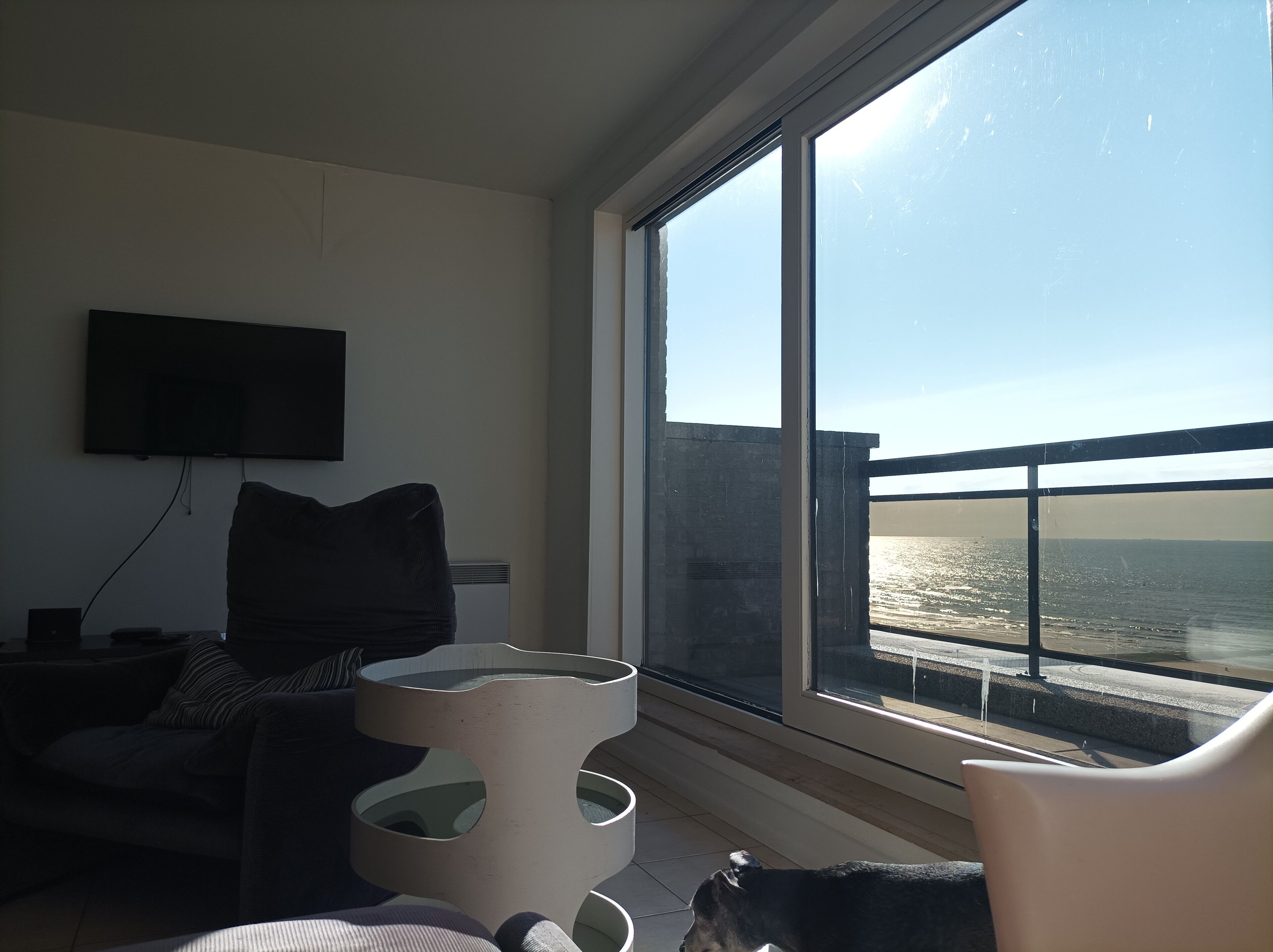 A modern living room with a large window overlooking a body of water, a flat-screen TV mounted on the wall, and a unique multi-tiered coffee table.