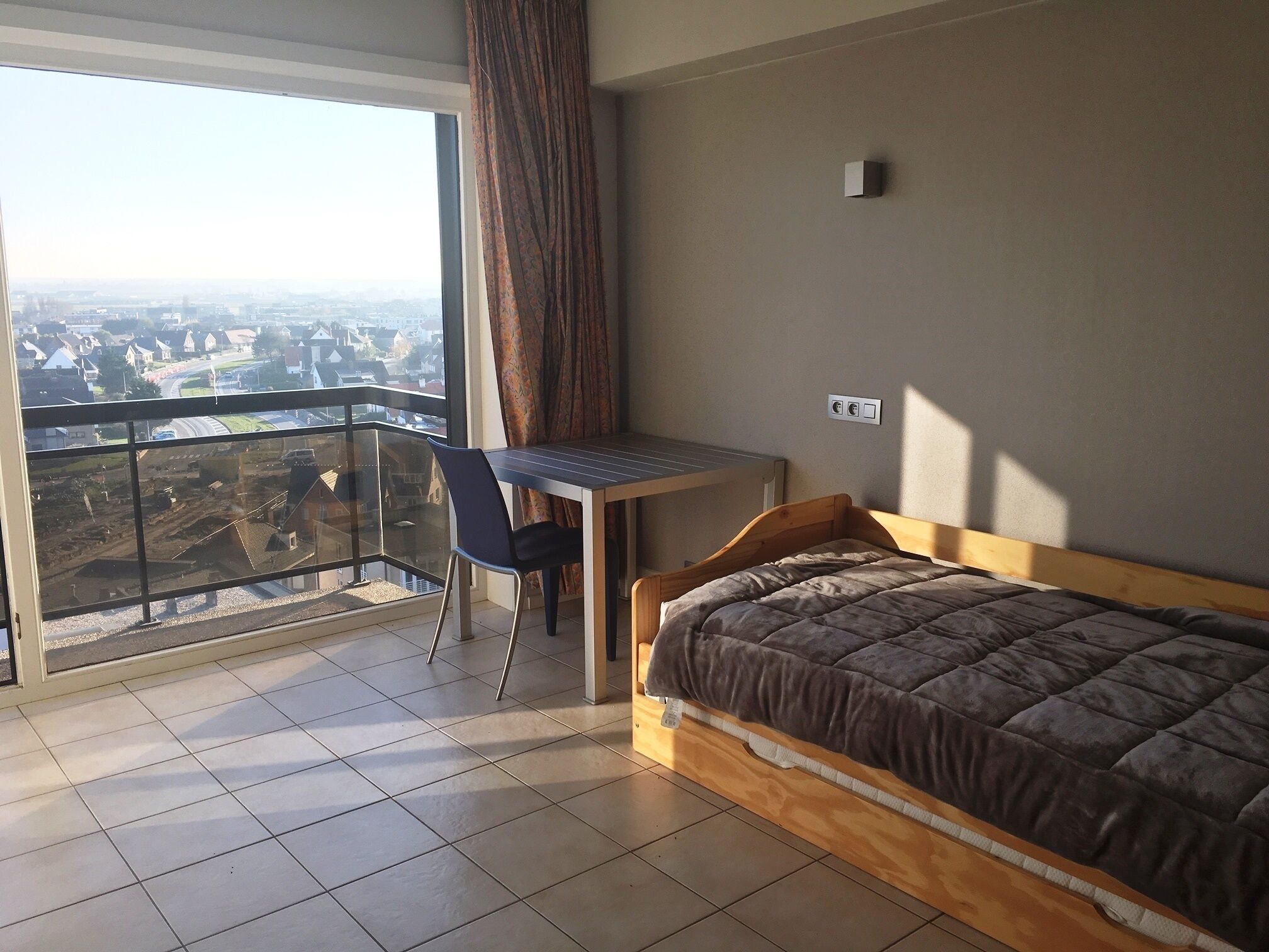 A bedroom with a bed, a desk, and a chair. There is a balcony with a view of the city.