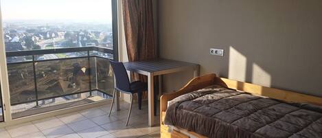 A bedroom with a bed, a desk, and a chair. There is a balcony with a view of the city.