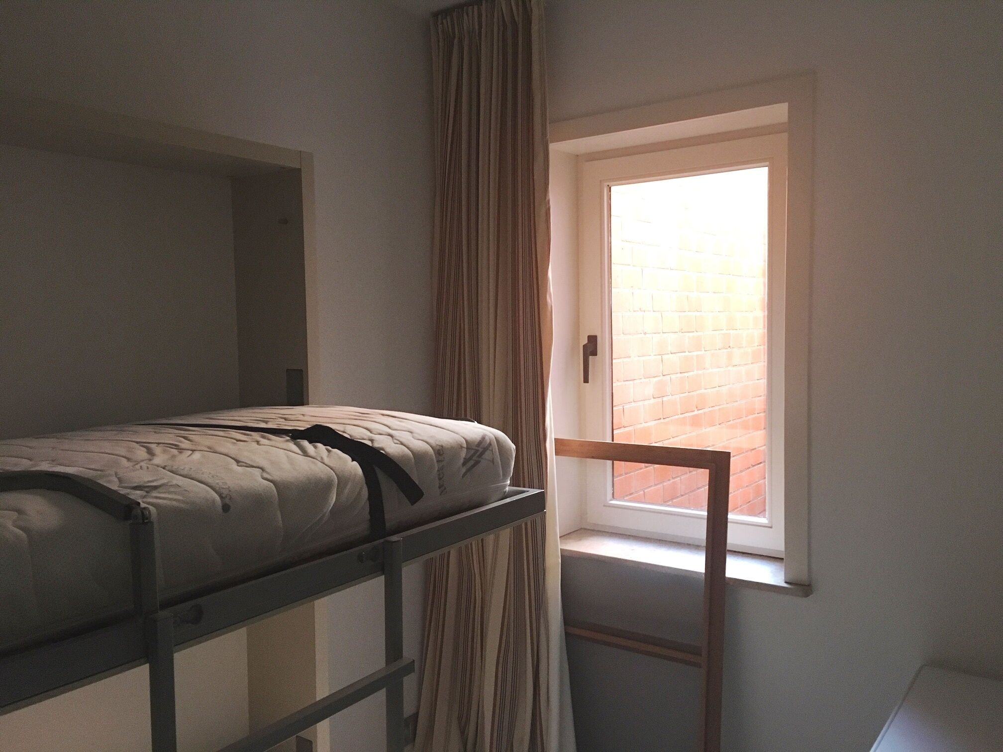 Une chambre de dortoir avec un lit superposé, une fenêtre avec des rideaux et un mur de briques visible à travers la fenêtre.