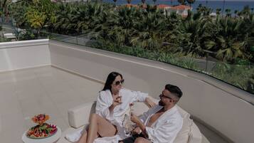 A couple relaxing on a white, round, cushioned seating area with a view of the ocean and palm trees.