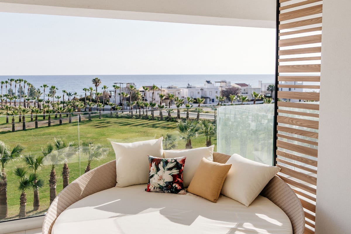 A round, wicker bed with a white mattress and beige pillows, positioned in a balcony with a view of a coastal landscape and palm trees.