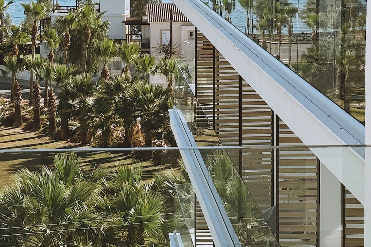 A modern building with a glass balcony overlooking a coastal area with palm trees and the sea.