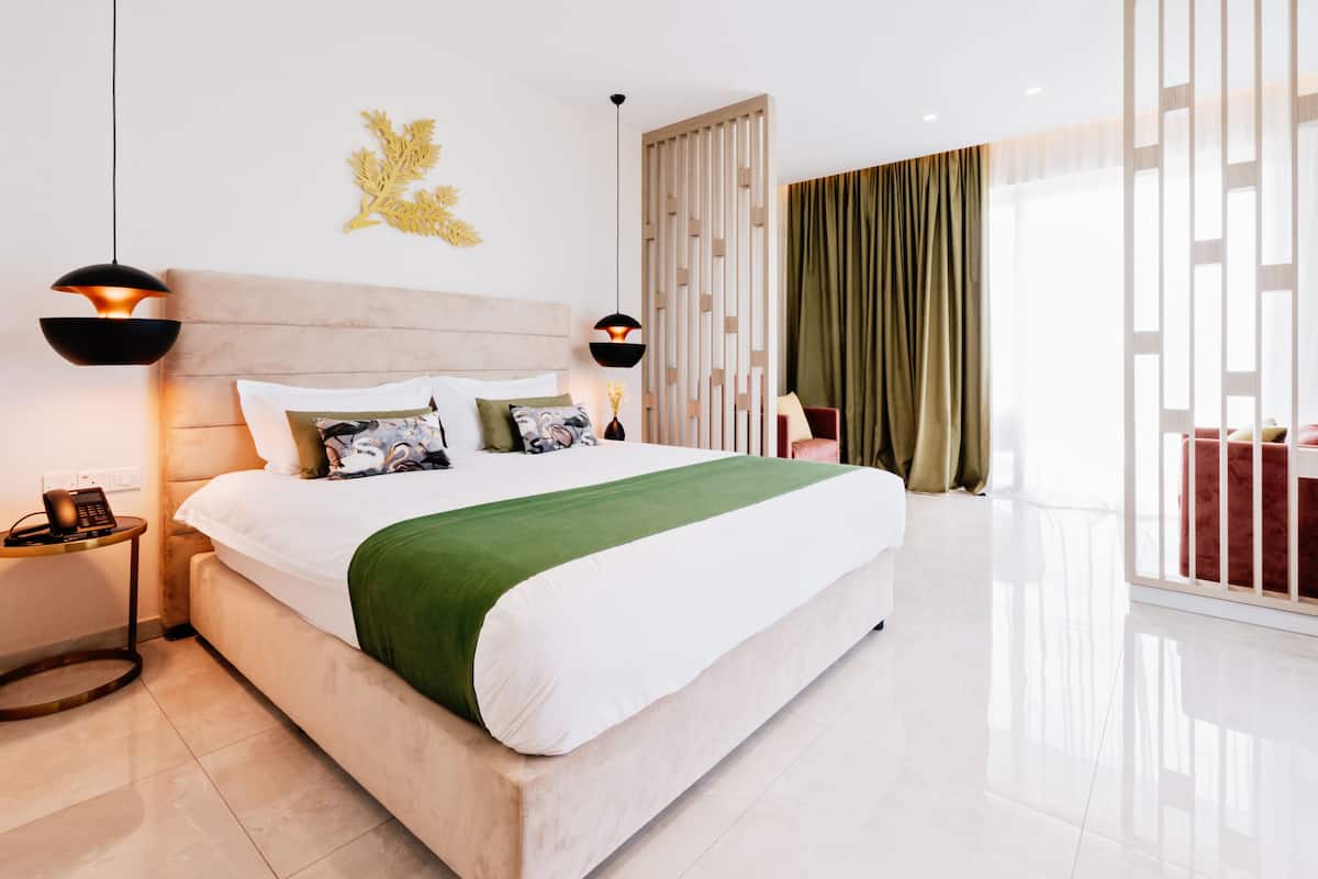 A modern hotel room with a large bed, two bedside lamps, a decorative wall piece, and a view of the outdoors through a large window with curtains.