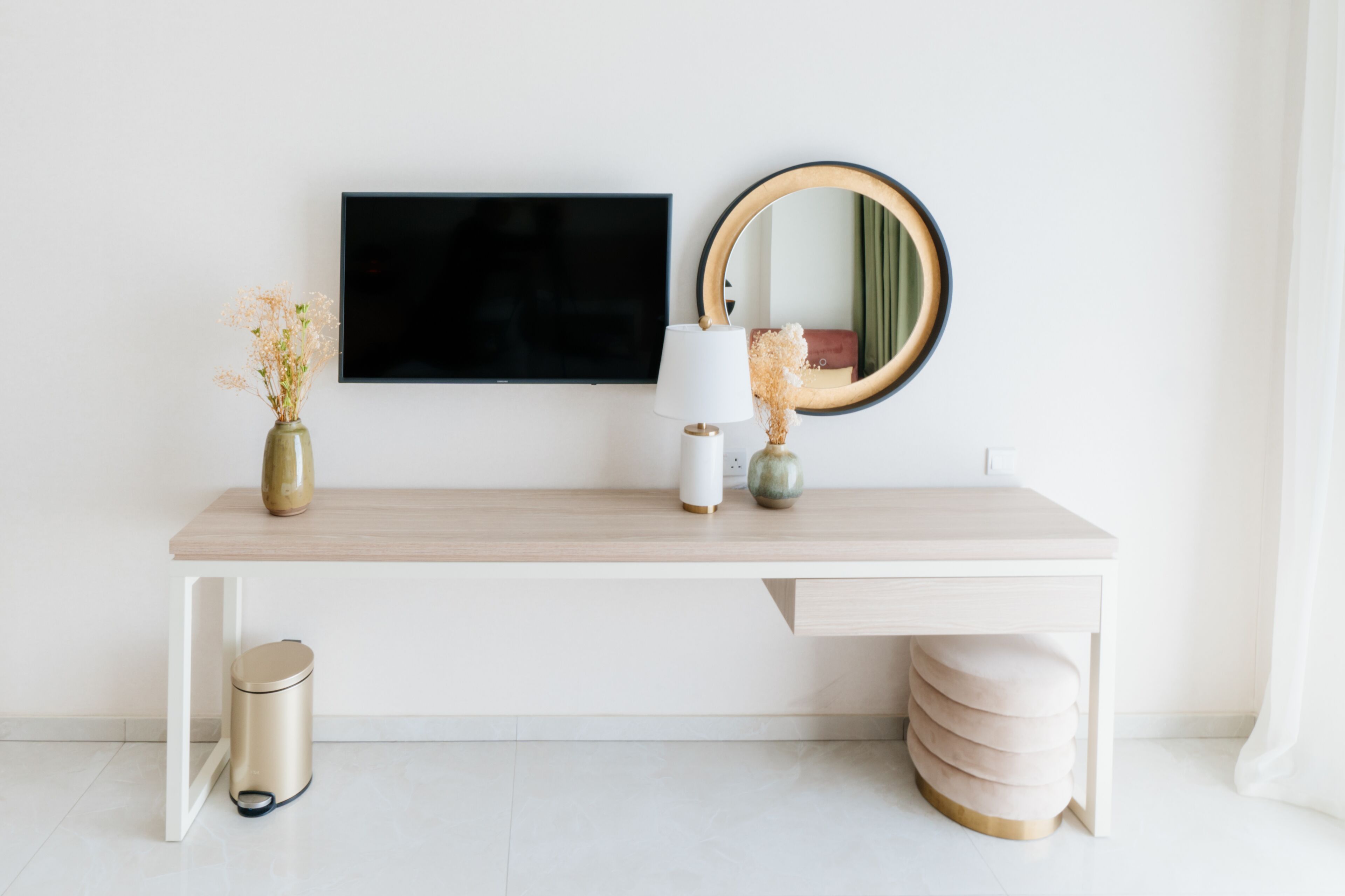 Une pièce au décor minimaliste, avec un bureau blanc, un téléviseur à écran plat, un miroir rond et une petite poubelle.