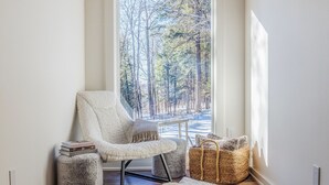 Une pièce chaleureuse avec une grande fenêtre, un fauteuil moelleux, un petit pouf, un panier en osier et un tapis duveteux.