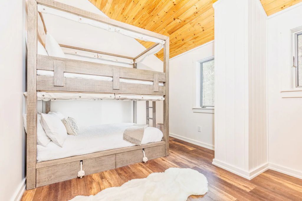 A bunk bed with a wooden frame and white bedding, a white rug on the floor, and a window with a view of the outdoors.