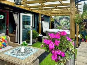 Ein überdachter Patio-Bereich mit einem Korbstuhl-Tisch, einem Blumentopf mit rosa Blüten und einem Whirlpool.