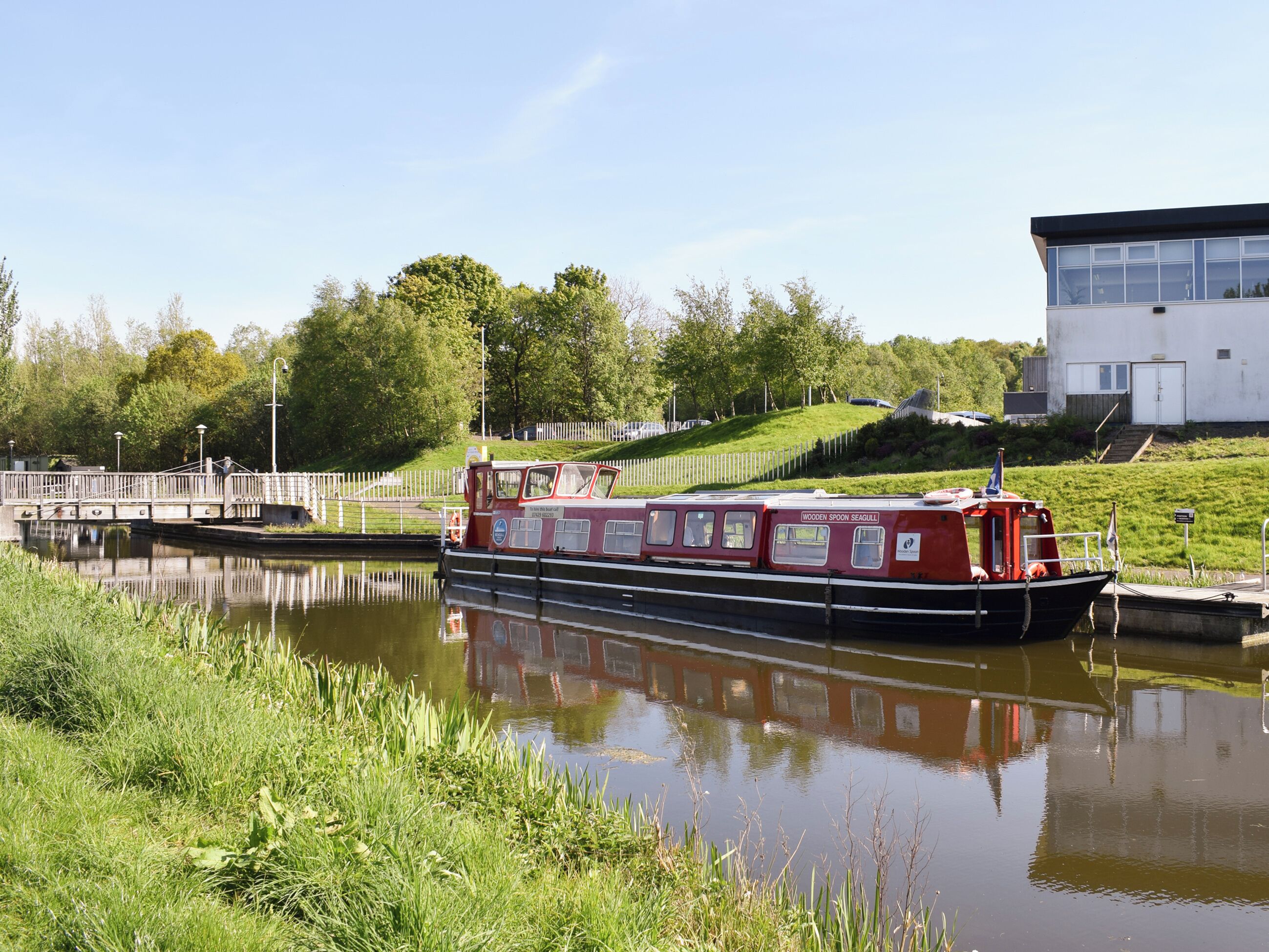 Un barco de canal amarrado en un canal, con una orilla cubierta de césped y un edificio blanco al fondo.