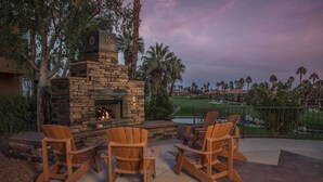 A stone fireplace with a fire, surrounded by wooden Adirondack chairs on a patio with a view of a golf course.