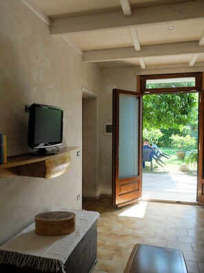 Ein Zimmer mit Fernseher auf einer Holzkammer, einem runden Holztisch und Blick auf einen Garten durch eine offene Tür.