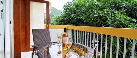 A round glass table with a wicker chair, a bottle of wine, and two wine glasses on a balcony with a yellow railing and green foliage in the background.