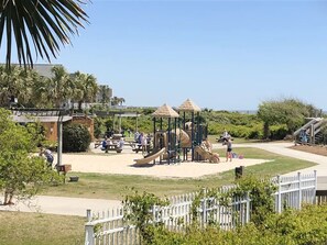 A playground with slides and climbing structures, surrounded by a white fence and palm trees.