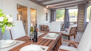 A wooden deck with a table set for dining, white cushioned chairs, and a view of the outdoors. - Mobile Home Pecten Exclusive 2 (Pakostane)