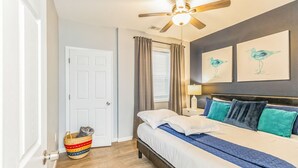 Une chambre à coucher avec un lit, un ventilateur de plafond, une fenêtre avec des rideaux et des œuvres d’art accrochées au mur.