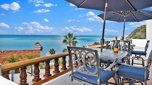 A balcony with a table and chairs, overlooking a coastal view with the sea and palm trees.